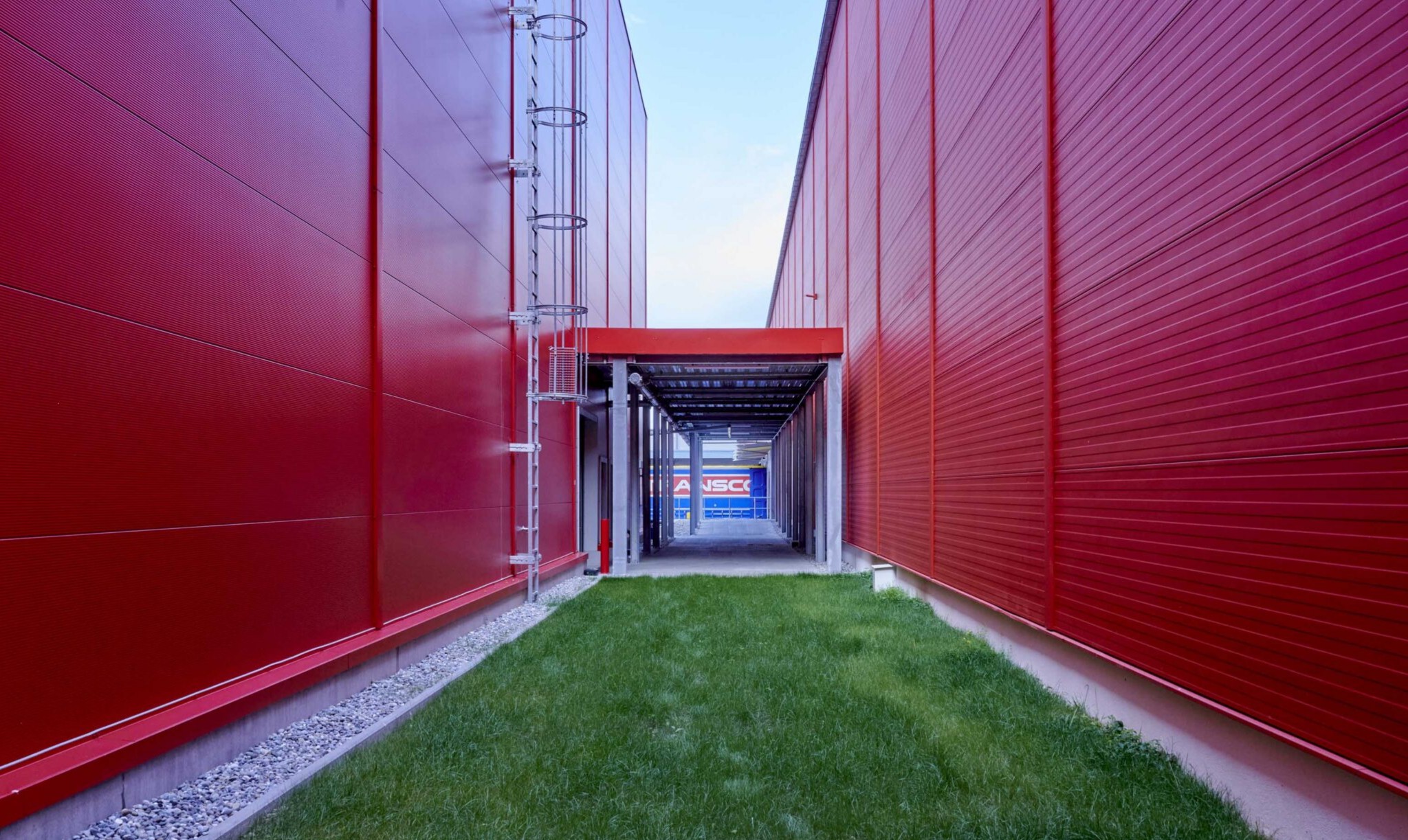 Schmaler Rasenstreifen zwischen zwei roten Lagerhallen mit Transco Lkw